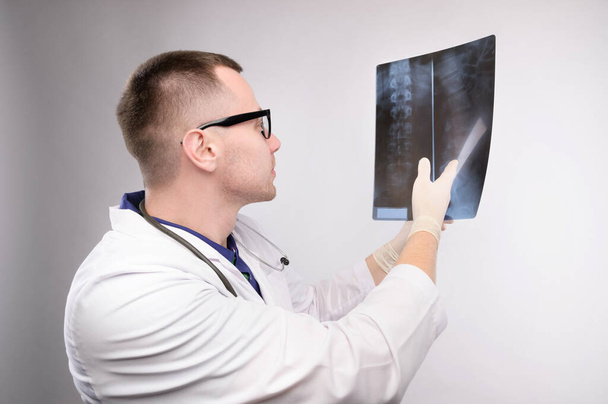 Young caucasian doctor a radiologist is looking at an x-ray. A doctor in a white coat with glasses with a stethoscope holds an x-ray in his hand - Photo, image