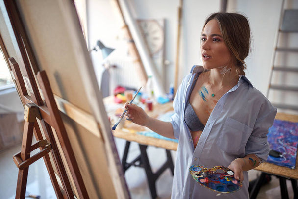 Close-up of a young hot and sexy female artist who is standing in front of the easel and working on a new painting in a relaxed atmosphere in her studio. Art, painting, studio - Foto, Imagem