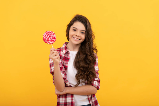 happy kid with long curly hair hold lollipop caramel candy on yellow background, sweet tooth. - Φωτογραφία, εικόνα