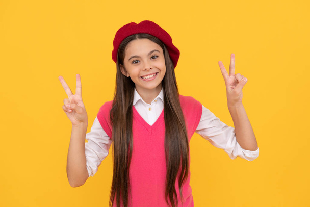 Ευτυχισμένος έφηβος κορίτσι σχολείο στα γαλλικά μπερέ δείχνουν χειρονομία ειρήνης σε κίτρινο φόντο, ειρήνη - Φωτογραφία, εικόνα
