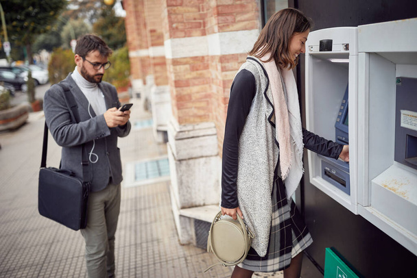 A young attractive woman is using ATM machine on the street in a relaxed manner. Walk, ATM, city - Fotoğraf, Görsel