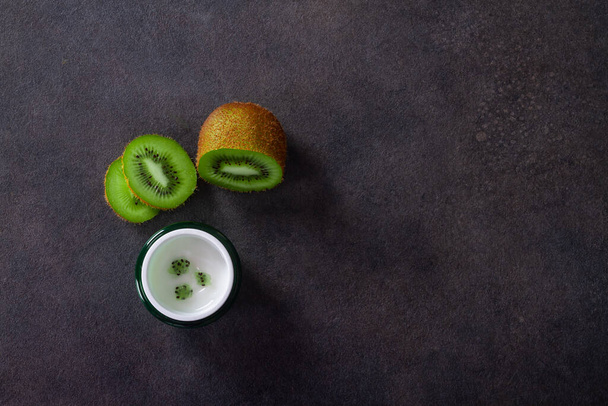 Cosmetic cream in a green jar with kiwi fruit on a black stone background. - Zdjęcie, obraz