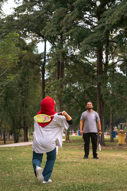 Азиатская семья играет в бадминтон в парке. Женщина в хиджабе, вид сзади. - Фото, изображение