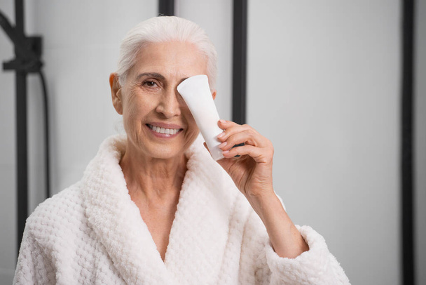 Portrait of smiling elderly woman in robe with bottle of cream in bathroom. Anti-aging cosmetics usage. Skincare at home. Cosmetology treatment - Φωτογραφία, εικόνα
