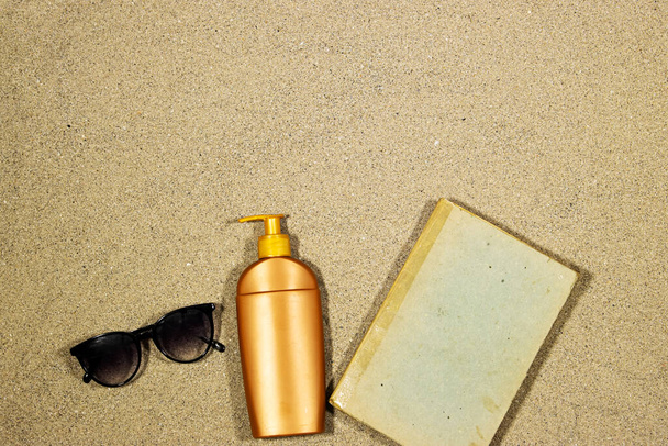 Composición laica plana con, crema protector solar con gafas de sol y libro viejo sobre arena. Fondo de vacaciones de verano con espacio de copia - Foto, Imagen