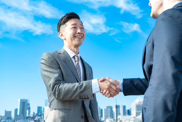 Businessmen shaking hands against the backdrop of the blue sky and buildings on fine day - Foto, afbeelding