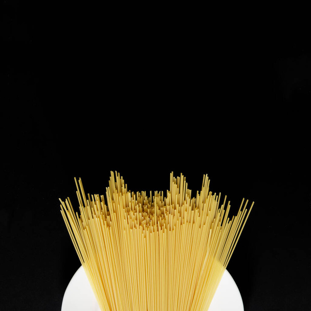 Bílý talíř a nevařené italské pšeničné špagety na černém pozadí. Pohled shora. Náměstí. Minimalismus. Volné místo pro nahrávání. Denní světlo - Fotografie, Obrázek