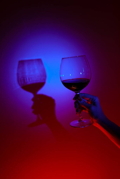 Großaufnahme einer Frau, die ein Glas Wein über die blaue Wand hält, neonblaues Licht an der Wand. - Foto, Bild
