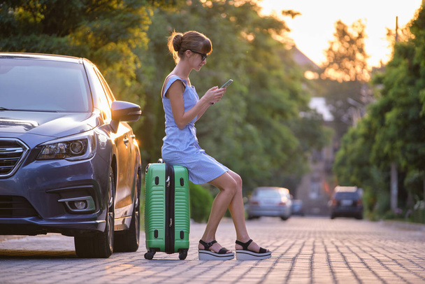Νεαρή γυναίκα κάθεται σε βαλίτσα κοντά στο όχημά της μιλώντας στο κινητό της τηλέφωνο σε δρόμο της πόλης το καλοκαίρι - Φωτογραφία, εικόνα