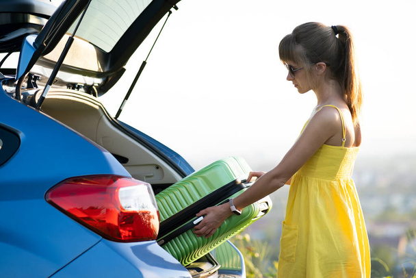 Young woman taking green suitcase from car trunk. Travel and vacations concept - Фото, изображение
