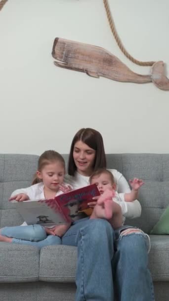 Una joven madre con un hijo y una hija pequeños lee un libro sentado en el sofá - Metraje, vídeo
