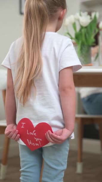 Ένα μικρό ξανθό κορίτσι κρατά μια κάρτα σε σχήμα καρδιάς για τη μητέρα της πίσω από την πλάτη της - Πλάνα, βίντεο