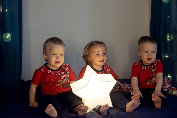 Тройняшки, сидящие на кровати, украшенные на Рождество. Трое милых маленьких детей в красных футболках - Фото, изображение