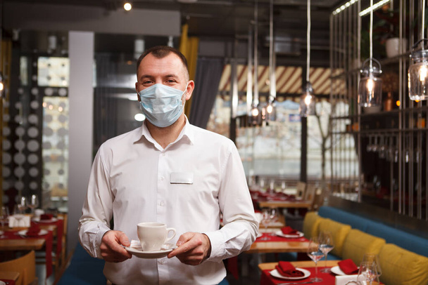 Male waiter wearing medical face mask, holding cup of coffee, copy space - Fotó, kép