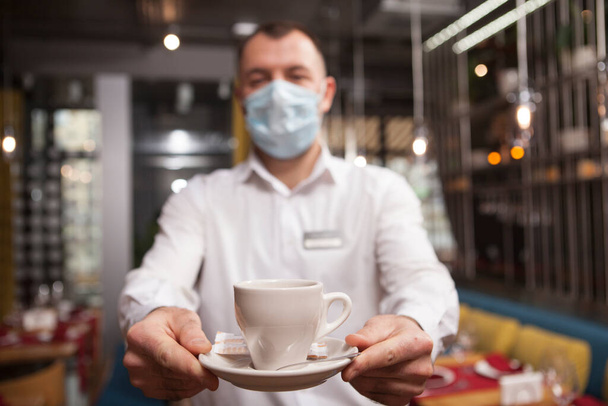 Selective focus on a coffee cup in hands of waiter wearing medical face mask - Foto, imagen