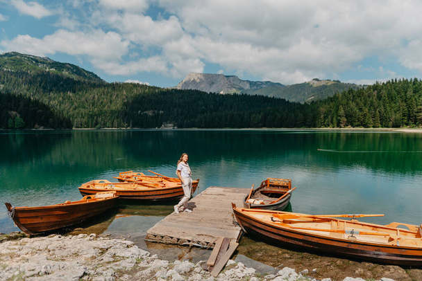 The girl looks at the Black Lake with fishing boats, forest and mountains in the Durmitor National Park of Montenegro. A tourist enjoys a magical view of the lake, a coniferous forest and a magical - Fotó, kép