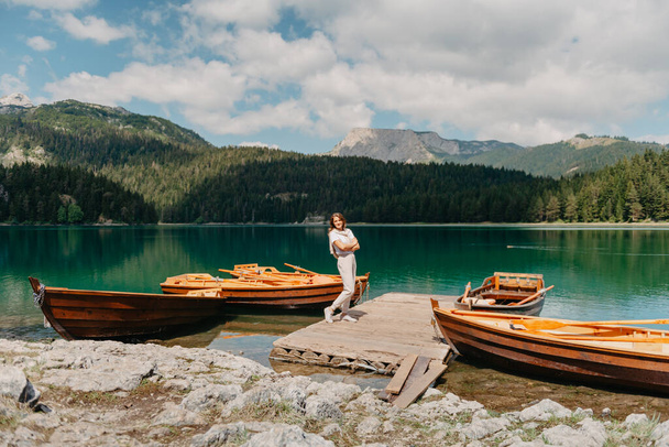 The girl looks at the Black Lake with fishing boats, forest and mountains in the Durmitor National Park of Montenegro. A tourist enjoys a magical view of the lake, a coniferous forest and a magical - Foto, Imagen