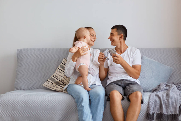 Horizontal shot of extremely happy young father and mother sitting on comfortable cough with their toddler daughter in living room, looking at each other and smiling, enjoying to spend time together. - 写真・画像