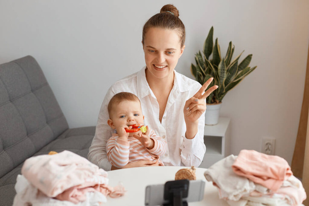 Smiling positive Caucasian mother sitting at table with toddler baby and using mobile phone on tripod for broadcasting livestream, showing v sign to smart phone camera, talking with subscribers. - Photo, image