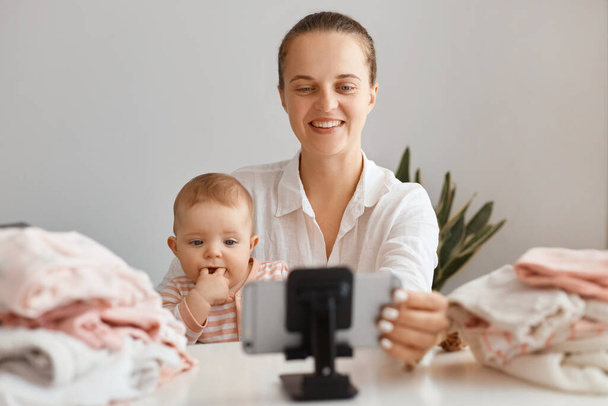 Positive woman sitting at table with toddler kid and recording video for her vlog, making content with her daughter, looking smiling at smart phone camera, livestreaming. - Fotoğraf, Görsel