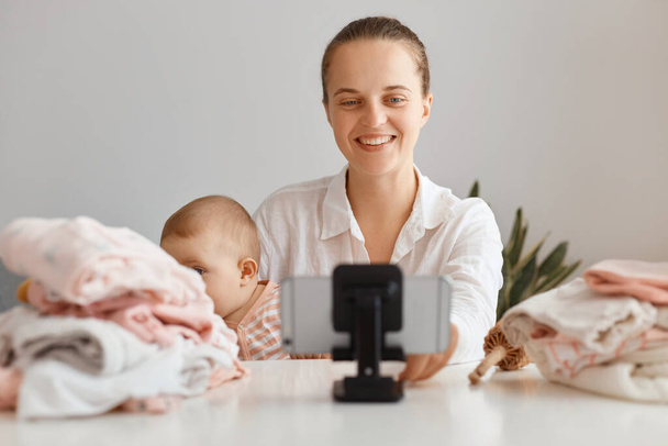 Image of young female blogger broadcasting livestream at home together with her infant daughter, filming content for her blog, looking smiling at phone camera, talking with subscribers. - Fotografie, Obrázek