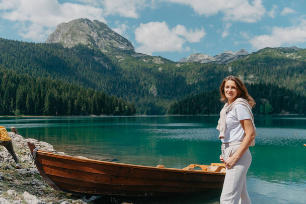 The girl looks at the Black Lake with fishing boats, forest and mountains in the Durmitor National Park of Montenegro. A tourist enjoys a magical view of the lake, a coniferous forest and a magical - Photo, Image
