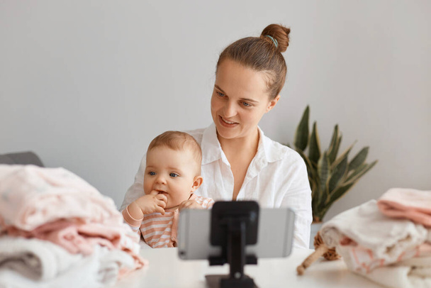Beautiful young adult woman blogger sitting at table with toddler kid and recording video for her vlog, using mobile phone on tripod, making content with her daughter. - Foto, afbeelding