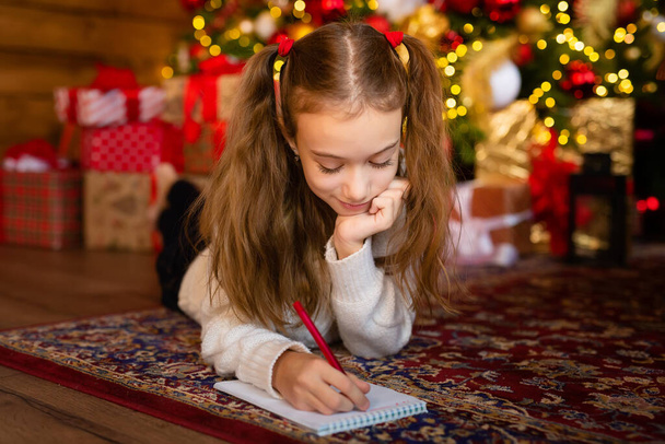 Una carta de niños a Santa en vísperas de un Feliz Año Nuevo. Plantilla, en blanco, postal. Una linda chica que escribe una carta a Santa cerca de un hermoso árbol de Navidad - Foto, imagen