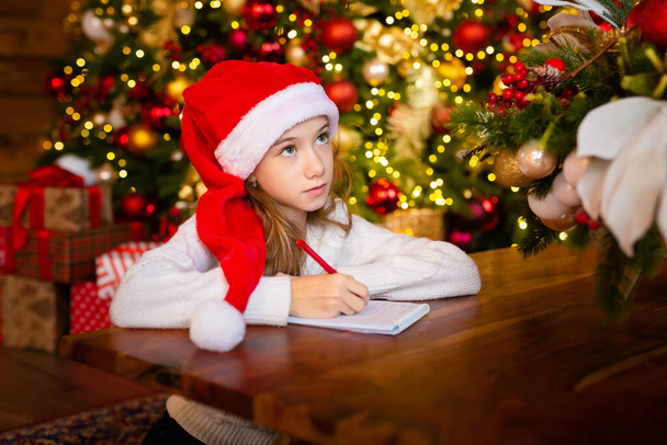 Boldog karácsonyt és boldog ünnepeket! Kislány télapó sapkában levelet ír Mikulás és álmok ajándék háttér szilveszteri fa beltérben. Üdvözlőlap, karácsonyi hangulat koncepció. - Fotó, kép