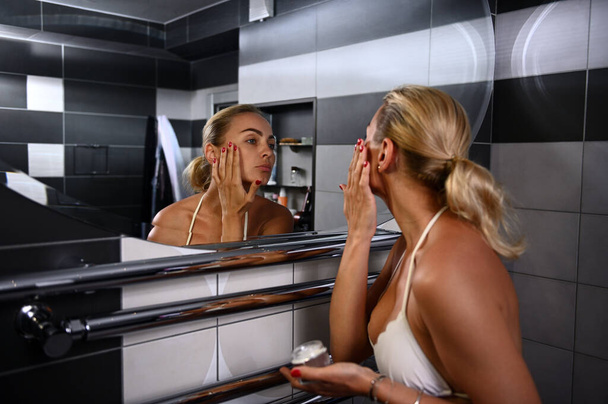 Очаровательная молодая кавказская блондинка в купальнике наносит увлажняющий крем, делает сглаживающий омолаживающий массаж лица, глядя на свое отражение в зеркале ванной комнаты - Фото, изображение