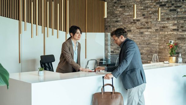 Female Hotel Receptionist Assisting Businessman for Checking In. Traveling During COVID-19: Business man with luggage passing passport and talking with concierge on hotel reception with sneeze guard protection.  - Foto, Imagen
