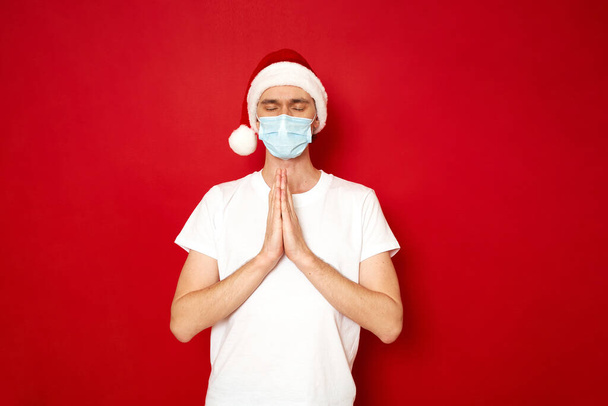 Modlitwa bożonarodzeniowa spokojnego człowieka o spełnienie pragnień, zdrowie bliskich, aby umieścić biały T-shirt w kapeluszu xmas, maska medyczna złożył ręce przed klatką piersiową z dłońmi skierowanymi do siebie - Zdjęcie, obraz