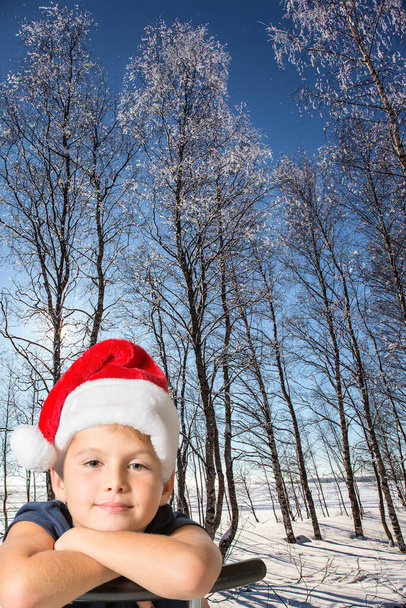 Christmas is coming. Frosty snowy sunny day in the winter forest. Deep snow covered the ground. Handsome boy in red Santa Claus hat smiles - Fotó, kép