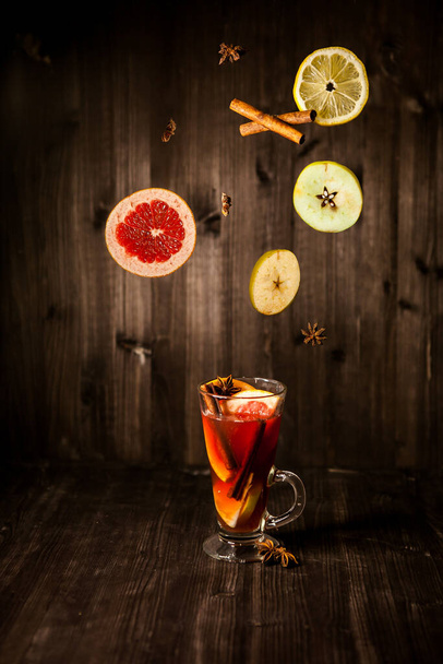 christmas mulled wine, in a transparent glass with fruit on a dark wooden background surrounded by flying spices and fruits - Foto, Imagem
