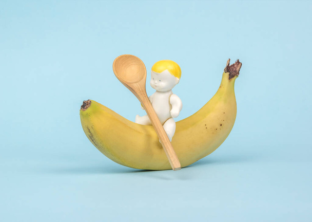 Una figura de un bebé sentado en un plátano con una cuchara de madera. Postre de plátano. El concepto de comida saludable para bebés. - Foto, imagen