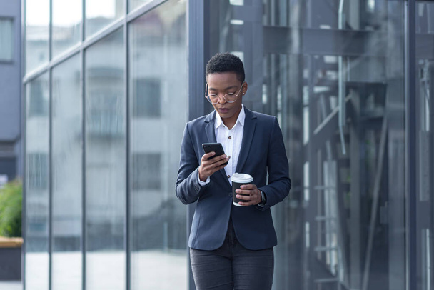 A confident African-American lady walks around the business district and uses the phone. Holding a cup of coffee, dressed in formal clothes. Attractive businesswoman browsing modern smartphone while communicating. applications - Foto, Imagem