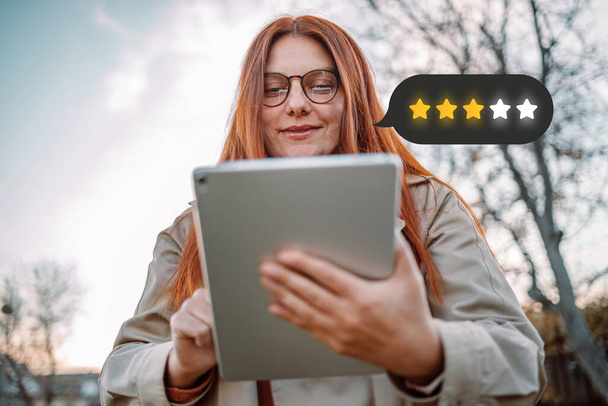 Jonge mooie blanke vrouw loopt op straat houdt moderne tablet notitieblok met pictogram 4-sterren symbool om de beoordeling van het bedrijf te verhogen en glimlacht vrolijk in de stad - Foto, afbeelding