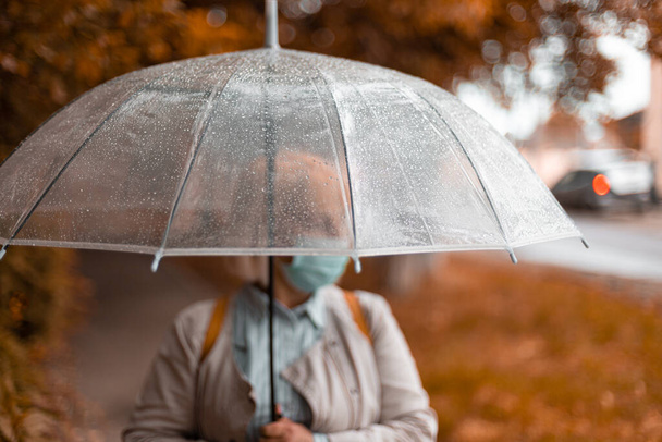 Retrato de mujer adulta con un sombrero blanco y una bufanda caliente bajo un paraguas transparente en un parque de otoño al aire libre, durante el día de lluvia fría - Foto, Imagen