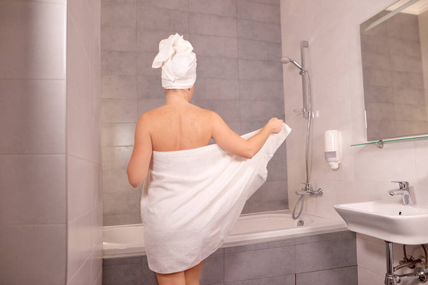 young woman stands with her back and takes off white towel to take a bath or shower. woman wipes herself off with a towel after shower - Foto, afbeelding