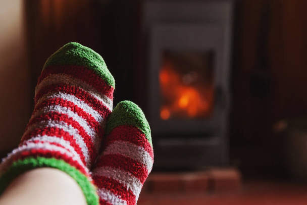 Feet legs in winter clothes wool socks at fireplace background. Woman sitting at home on winter or autumn evening relaxing and warming up. Winter and cold weather concept. Hygge Christmas eve. - Foto, immagini