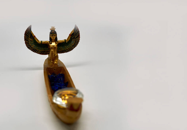 エジプトのお土産を飾りました。翼のある女神イシスと黄金のボート。イシスはヘッドギアを失った。船には青いガラスの石とダイヤモンドの形をしたガラスのクリスタルがあります。.                               - 写真・画像