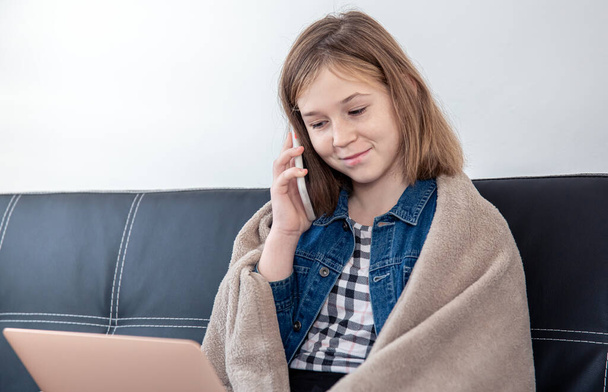 Una adolescente está sentada en el sofá en un portátil y hablando por teléfono. - Foto, imagen