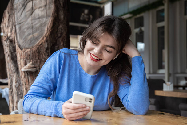 Молода жінка в бездротових навушниках використовує телефон, сидячи в кафе
. - Фото, зображення