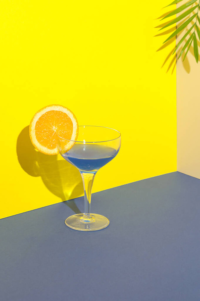 Blauwe cocktail met een schijfje sinaasappel op de rand van het glas op een combinatie van blauw geel en crème ondergrond. Palmblad op de achtergrond. Zomer concept drank sap voor verfrissing, feesten... - Foto, afbeelding