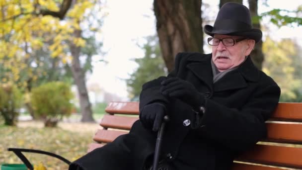 Oudere eenzame grootvader zittend op een bankje met een stok in het herfststadspark. - Video