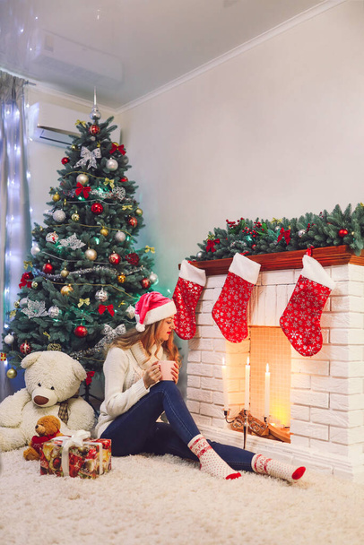 Giovane signora in maglione bianco seduta con caminetto pulito tazza. albero di Capodanno e decorazione domestica. Candele e orsacchiotto, i regali sono in giro. Concetto di vacanze di Natale. Atmosfera calma e pacifica - Foto, immagini