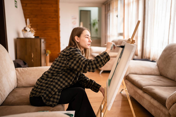 One young beautiful caucasian woman artist sitting on the bed sofa at home painting on the canvas holding paintbrush art and creativity professional or hobby concept real people side view copy space - 写真・画像