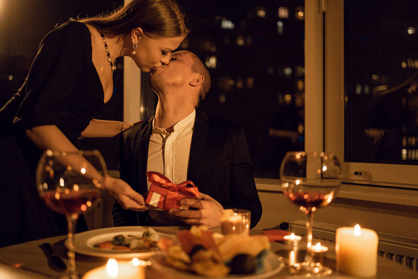 Passionate kiss at dinner on a date, a woman gives a man a gift box on Valentine's Day, a romantic dinner in a restaurant, an intimate date of a couple, flirting in private - Photo, image