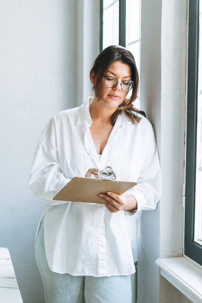 Giovane donna bruna sorridente plus size in camicia bianca vicino alla finestra con penna e documenti in mano nel luminoso ufficio moderno - Foto, immagini