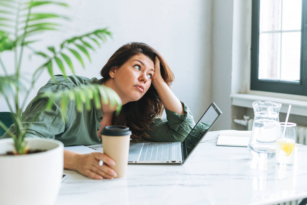 młody myślenie nieszczęśliwy brunetka kobieta plus rozmiar pracy w laptop na stole z dom roślin w jasny nowoczesny biuro - Zdjęcie, obraz